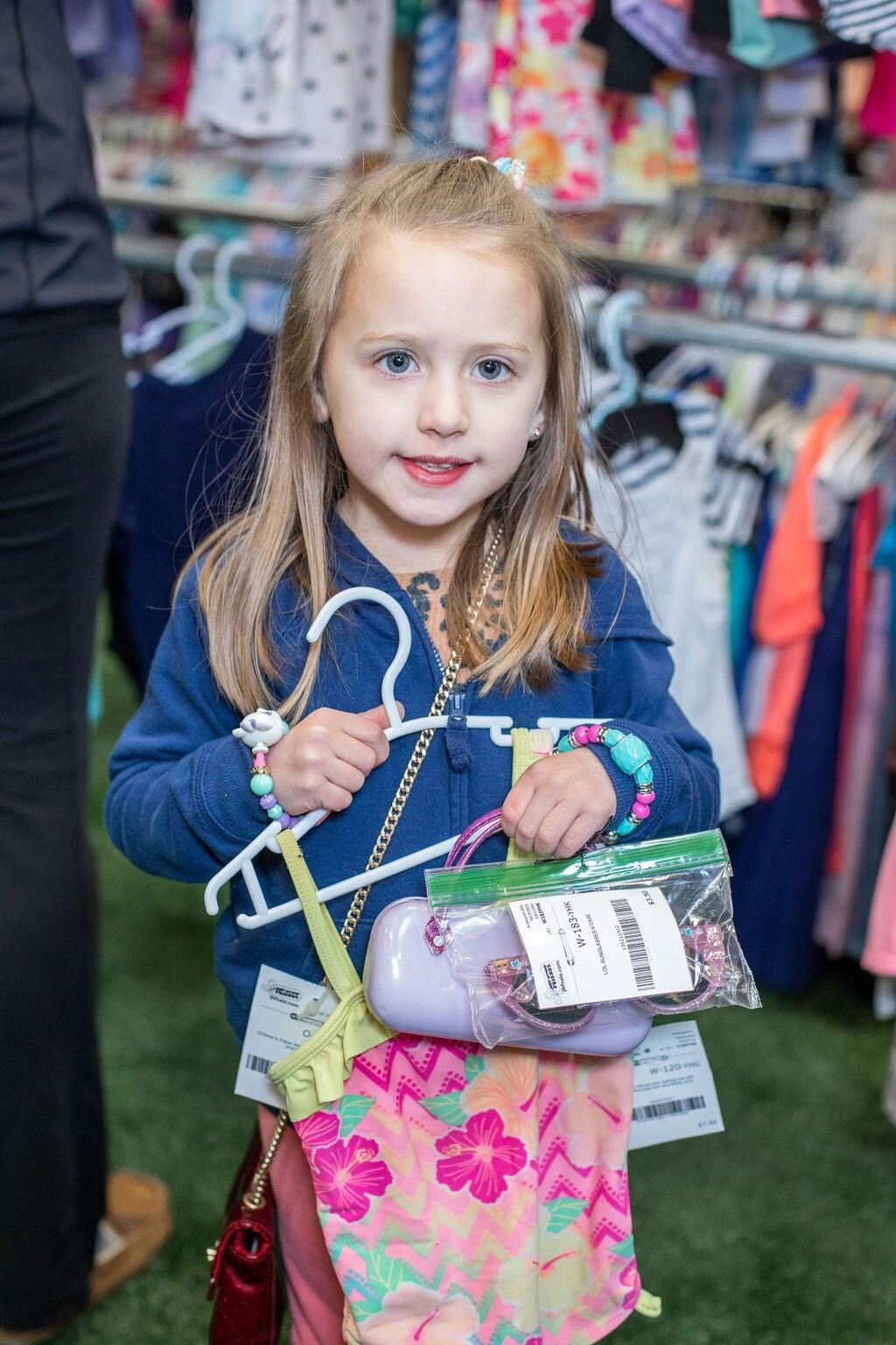 A woman kneels in front of racks of boys clothing. She is hold hangers full of boys clothing in both hands.
