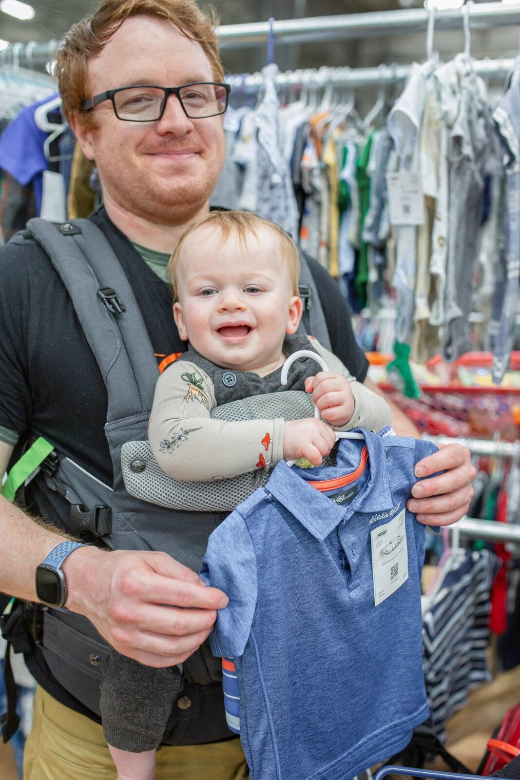 A woman is carrying her baby in a front carrier and holding a purple JBF bag in one hand and a hanger with a pair of demin overall shorts in her other hand. She is standing next to a rack of clothing for sale.