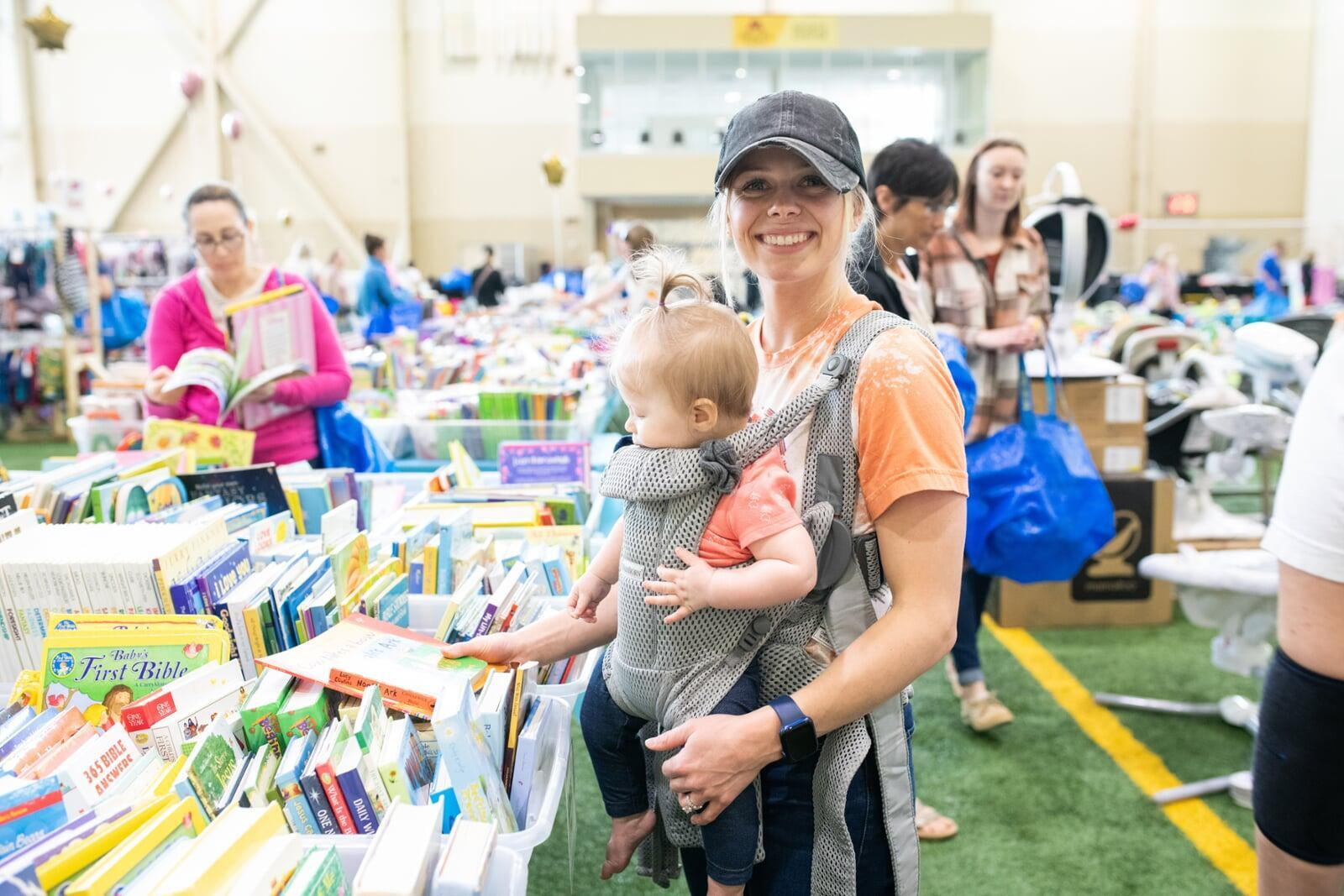 Couple shopping at the JBF sale. Mom has a baby in a front carrier and dad is holding up an activity walk-behind toy.