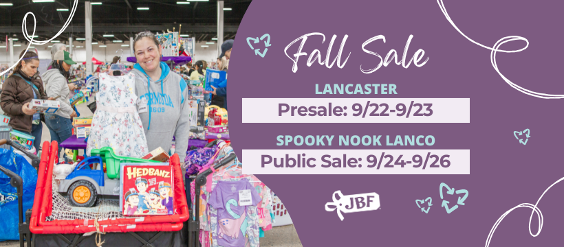 Two ladies smile as the hold up hangers with clothing on them. They are both standing next to a rack full of clothing for sale.