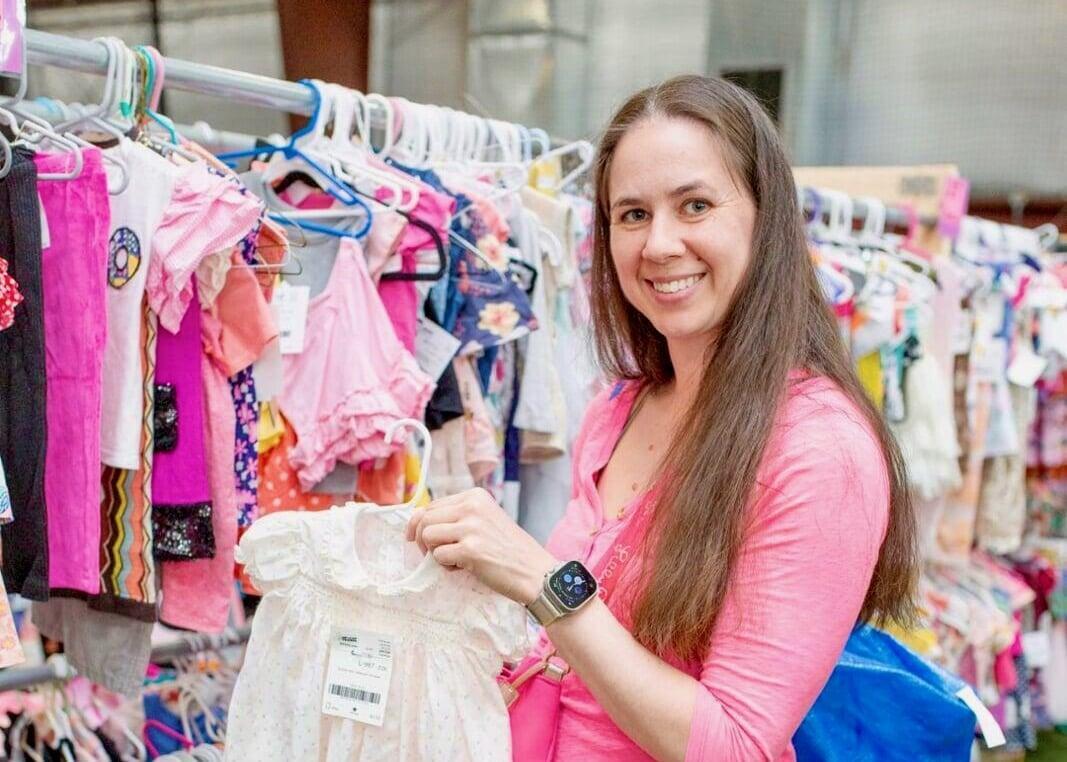 Two friends smile and laugh and are holding toddler girls clothing on hangers. They are standing between racks of girls clothing.
