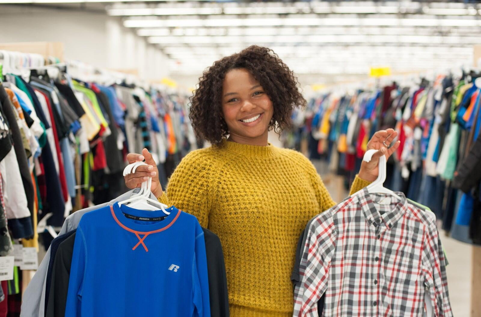 Mom is wearing a gold sweater and holding up two items of boys clothing on hangers.