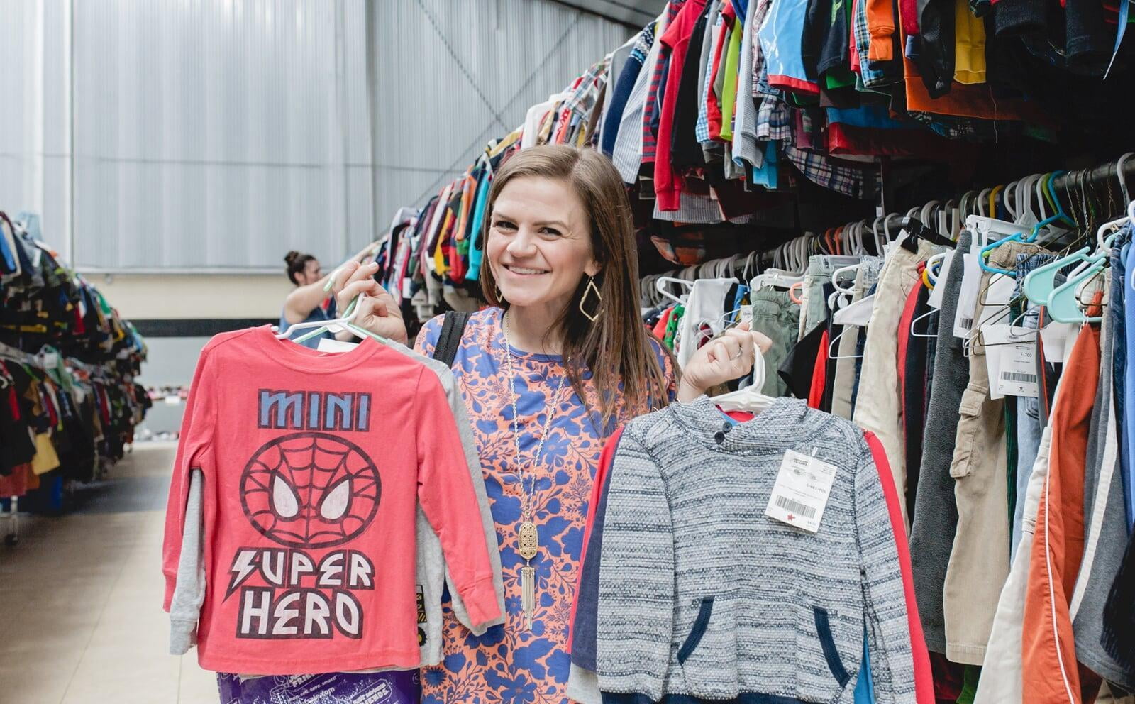 A woman kneels in front of racks of boys clothing. She is hold hangers full of boys clothing in both hands.