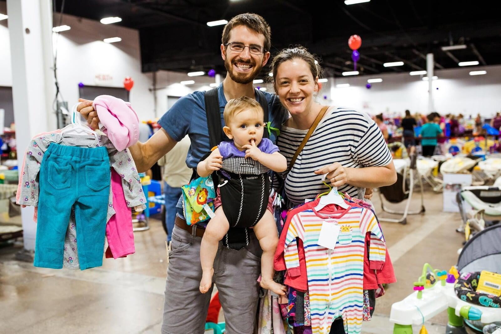 Young couple shopping with their baby.  Dad has daughter in a front pack carrier and is holding up girl's clothing.  Mother is also holding girl's clothing.